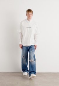 Balta trumpomis rankovėmis marškiniai su Calvin Klein logo, dėvimi virš balto ilgomis rankovėmis marškiniai; šviesiai mėlyni nusidėvėję plačios kojomis džinsai; balti sportbačiai.