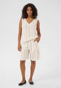 Débardeur sans manches beige clair et shorts assortis à rayures verticales ; les shorts ont des poches ; le modèle porte des ballerines noires, cheveux en boucles lâches.