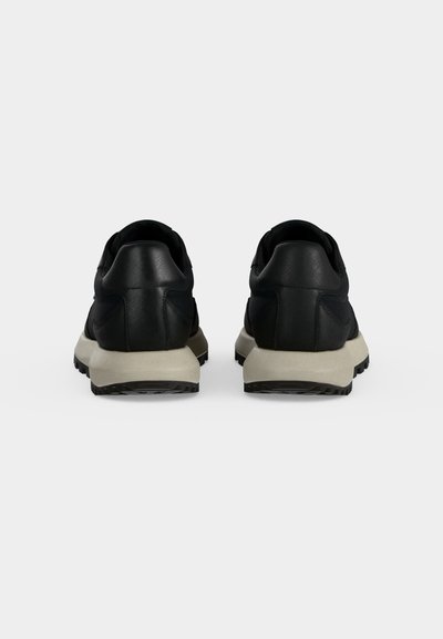 Paire de baskets basses noires avec semelles beiges vues de dos sur un fond gris clair uni.