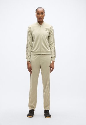 Tuta beige chiaro in tessuto liscio, composta da una giacca con zip e colletto, con dettagli a righe sulle maniche, abbinata a pantaloni coordinati.