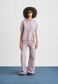 Ανοιχτό μωβ φούτερ με κουκούλα και τσέπη μπροστά, με λογότυπο. Συνδυάζεται με αντίστοιχο παντελόνι φαρδιάς γραμμής. Λείο, απαλό ύφασμα.