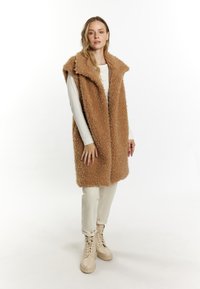 Beige nepbont vest met een kraag, open voorkant en zijzakken, gedragen over een lichtgekleurde langemouwenhemd, gecombineerd met crèmekleurige broek en laarzen.