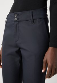Navy trousers with a smooth texture, featuring a flat waistband, two buttons, and side pockets. Close-up of waist and hand details.