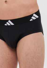 Schwarze Herrenunterwäsche mit einem enganliegenden Schnitt, ausgestattet mit einem breiten Bund, der mit drei weißen diagonalen Streifen verziert ist. Der Stoff wirkt glatt mit einem leichten Glanz.