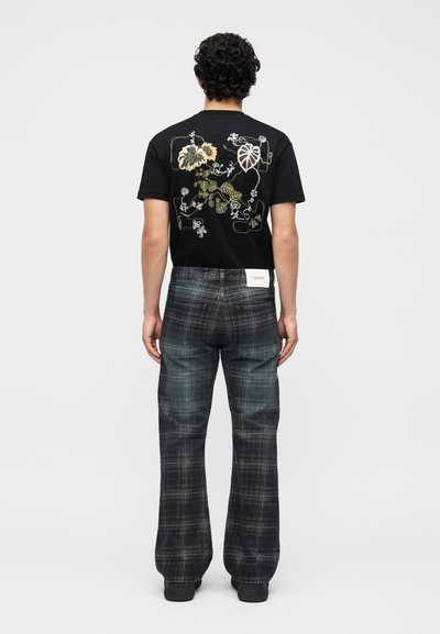 Άνδρας που φοράει μαύρο μπλουζάκι με κεντητό βοτανικό σχέδιο και σκούρο καρό παντελόνι με φαρδιά μπατζάκια, στέκεται γυρισμένος με την πλάτη σε μονόχρωμο φόντο.