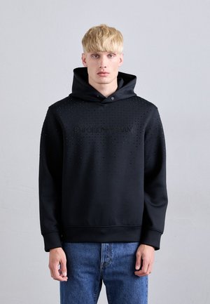 Schwarzer Hoodie mit strukturierter Oberfläche und Nieten-Details. Auf der Vorderseite steht "EMPORIO ARMANI" in Schwarz, kombiniert mit blauen Jeans.