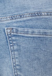 Tecido de denim azul claro com um bolso traseiro de borda reta e costura visível. A textura está ligeiramente desbotada com um acabamento suave.