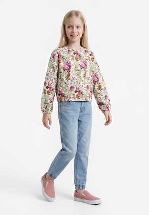 Mladé dievča sa usmieva, nosí kvetovaný dlhý rukáv top, svetlomodré rifle s elastickými manžetami a ružové slip-on tenisky, stojí na bielom pozadí.