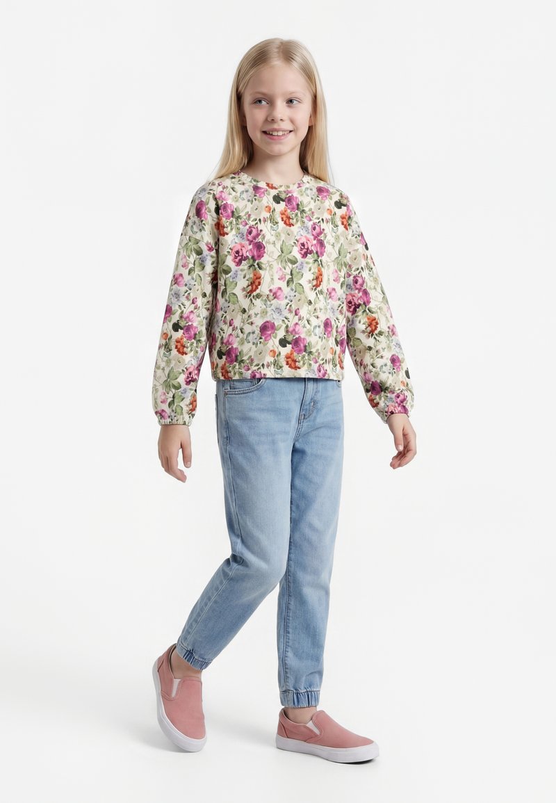 Joven niña sonriendo, vestida con una camiseta de manga larga floral, vaqueros azul claro con puños elásticos y zapatillas rosas sin cordones, de pie sobre fondo blanco.