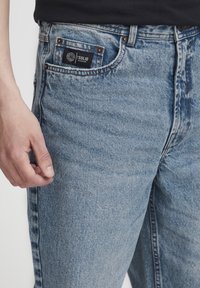 Hele sinised lahtise lõikega teksapüksid. Disainis on eesmine tasku musta sildiga, vasksed naelad ja nähtavad õmblused.