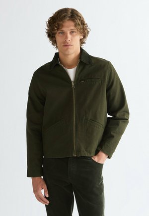 Mann mit lockigem Haar trägt eine dunkelgrüne Zip-Jacke über einem weißen T-Shirt und posiert mit einer Hand in der Hosentasche vor einem schlichten Hintergrund.