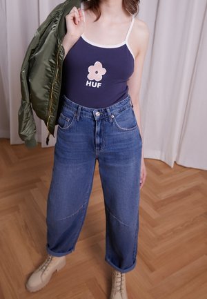 Person iført navy tanktop med blomst og teksten "HUF", højttaljede blå jeans, beige støvler, holder grøn jakke over skulderen.
