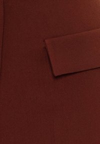 Hnědý vlněný kabát zblízka ukazuje obdélníkovou náprsní kapsu s klopou na hladké látce.