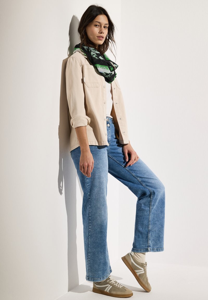 Beige button-up skjorte over en hvid top, parret med lyseblå flagrende jeans og beige sneakers. Et sort og grønt mønstret tørklæde fremhæver looket.