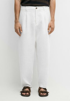 Mann trägt lockere weiße Leinenhose und braune Sandalen mit Doppelriemen, steht vor weißem Hintergrund.