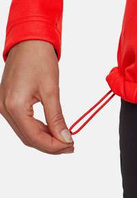 Hand justiert roten Kordelzug am Saum einer roten Jacke, trägt schwarze Hose vor weißem Hintergrund.