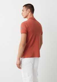 Polo shirt color ruggine in cotone con maniche corte, dotato di uno spacco laterale e di un logo circolare sulla manica sinistra. Indossato con pantaloni cargo bianchi.