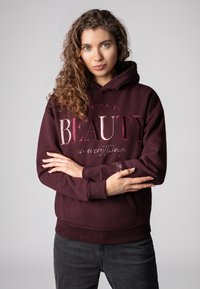 Bordeauxfarbener Kapuzenpullover mit einem Frontdruck, der "SCHÖNHEIT" in verschiedenen Pinktönen zeigt. Verfügt über eine Kapuze mit Kordelzug und gerippte Bündchen.