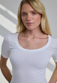 Blondynka o niebieskich oczach w białej koszulce z krótkim rękawem i dekoltem typu scoop, stojąca na tle jasnej ściany z cieniami.