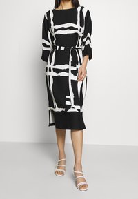 Abito nero con motivo astratto bianco, maniche a tre quarti, vita con cintura, lunghezza al ginocchio con spacco laterale, abbinato a sandali bianchi con cintura.