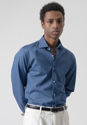 Joven con cabello rizado corto, vistiendo una camisa azul de botones metida dentro de pantalones blancos con un cinturón negro, posando frente a un fondo liso.