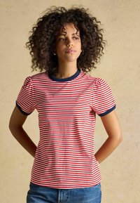 Femme aux cheveux bouclés portant un t-shirt rayé rouge et blanc avec des bordures bleu marine, debout les mains derrière le dos devant un fond beige.