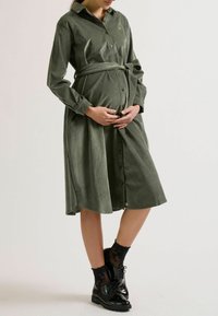 Schwangere Frau in einem grünen Hemdkleid, die sanft ihren Bauch hält, trägt schwarze Schuhe und gemusterte Socken, vor einem neutralen Hintergrund.