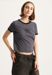Grå beskåret t-shirt med sorte ringer detaljer, der har et grafisk print p�å forsiden. Stoffet ser blødt ud med en afslappet pasform.
