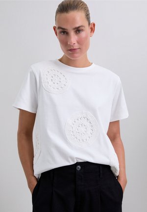 Femme avec les cheveux plaqués en arrière, portant un t-shirt blanc avec des motifs en dentelle circulaires et un pantalon noir taille haute, les mains dans les poches.
