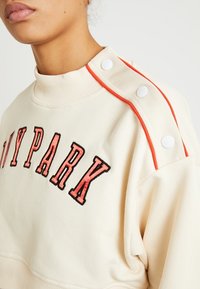 Krémová mikina s výrazným červeno-černým nápisem „IVYPARK“, žebrovaným výstřihem a druky s oranžovými akcenty na rameni.