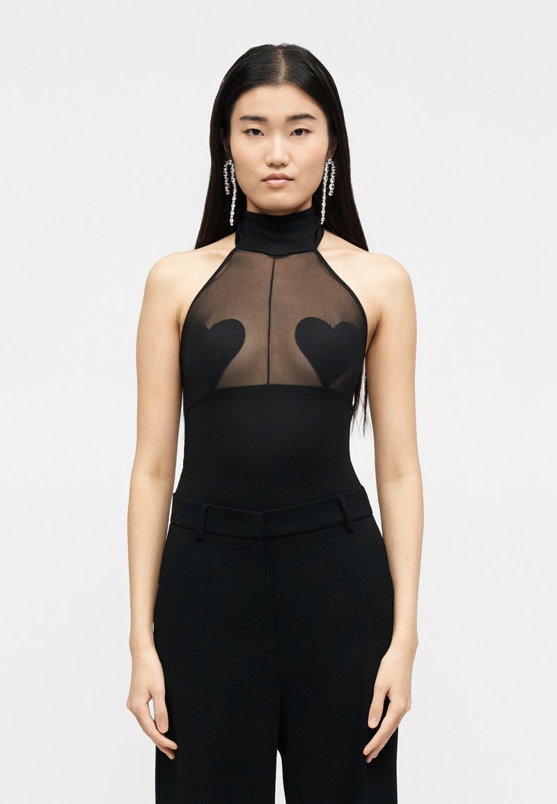 Jeune femme aux longs cheveux foncés portant un top noir sans manches avec une section supérieure transparente et des patches en forme de cœur, associée à un pantalon noir et des boucles d'oreilles pendantes.