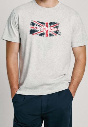 Hombre con camiseta gris claro con la Union Jack descolorida y texto "Pepe Jeans London", combinado con pantalones azul oscuro, manos en los bolsillos.