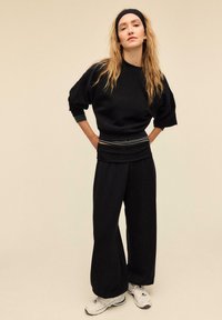 Sort oversized sweatshirt med puffede ærmer og stribede manchetter, kombineret med løse sorte bukser og hvide sneakers, mod en neutral baggrund.