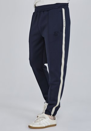 Marineblaue Jogginghose mit Seitentaschen, ausgestattet mit weißen Streifen entlang der Seiten und einem gestickten Logo am linken Oberschenkel. Weicher Stoff, schmal zulaufende Passform.