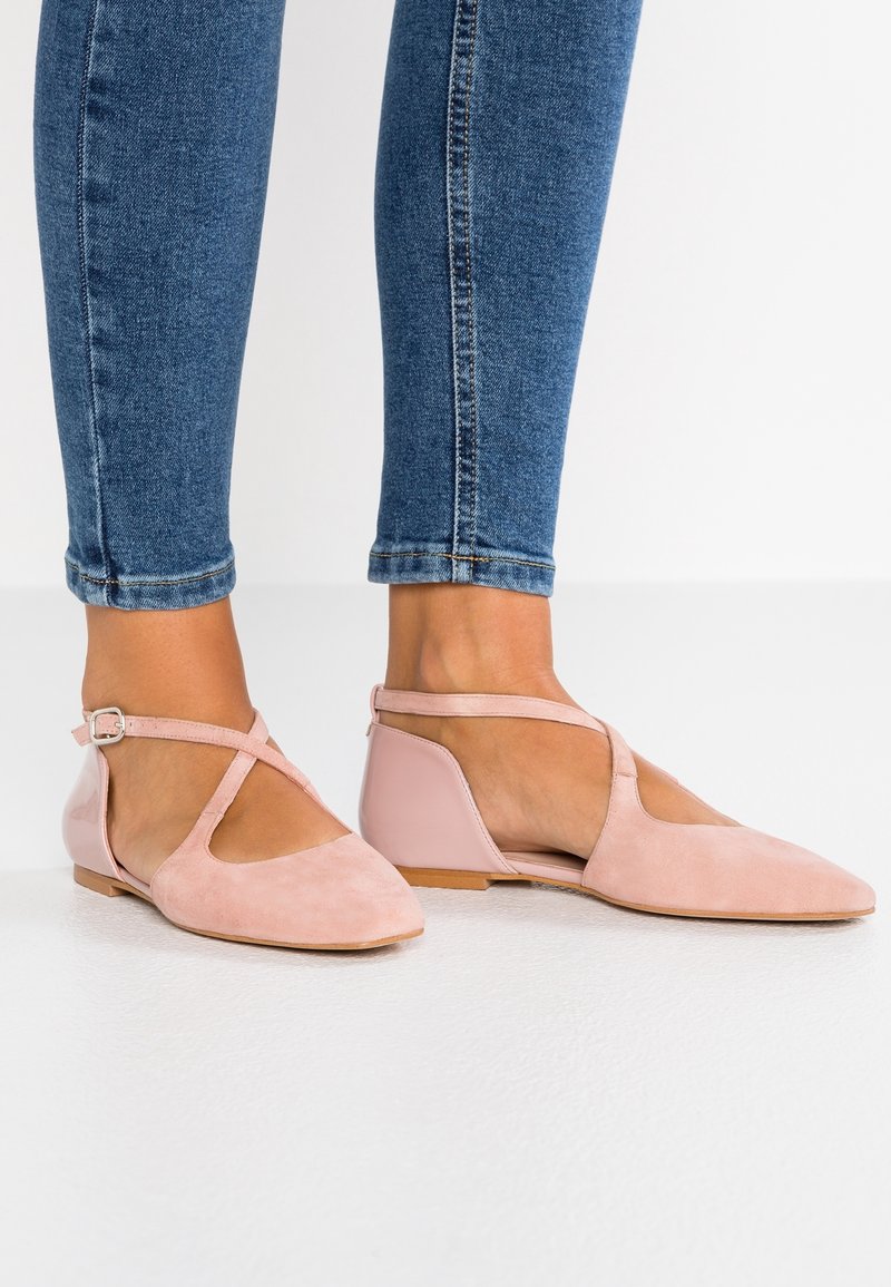 Pieds chaussés de chaussures plates en daim rose pâle avec des brides croisées autour de la cheville, assorties à un jean skinny bleu sur un fond blanc uni.