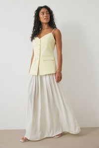 Kollane kohandatud vest nööpidega, koos voolava valge maxi-seelikuga. Riietusel on õhukesed spaghetti-rihmad ja sile tekstuur.