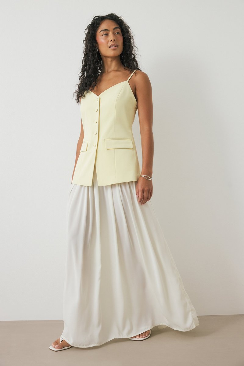 Kollane kohandatud vest nööpidega, koos voolava valge maxi-seelikuga. Riietusel on õhukesed spaghetti-rihmad ja sile tekstuur.