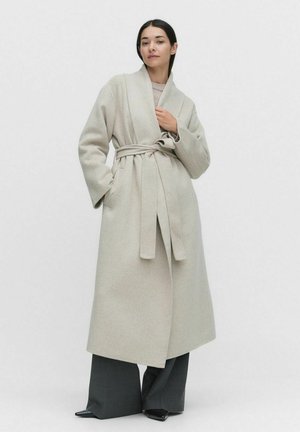 Ljusgrå ullkappa med omslagdesign, bälte vid midjan och vida ärmar, kombinerad med mörka, vida byxor och spetsiga stövlar.