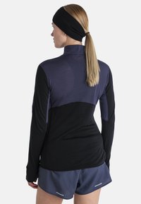 Langärmliges Sportoberteil in Schwarz mit einem oberen Bereich in Lila, ausgestattet mit einem Stehkragen und enganliegenden Ärmeln, kombiniert mit marineblauen Shorts.