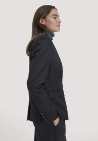 Donkere getailleerde blazer van soepel stof. Beschikt over een enkelgeknoopt ontwerp, inkepingen op de revers en manchetknopen met vier knopen, gestyled met een coltrui.