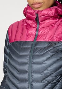 Eristetty takki vaaleanpunaisella ja harmaalla värimaailmalla; siinä on korkea kaulus, vetoketjukiinnitys, vaakasuora chevron-tikkaus ja musta logo.