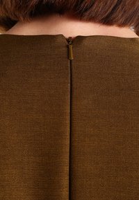 Hnědá texturovaná látka s viditelným zadním zipem. Stahovák zipu je lesklé zlaté barvy, což zvýrazňuje detail zapínání oděvu.