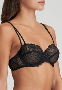 Schwarzer Spitzen-BH mit transparenten Einsätzen, gepolsterten Cups und gewellten Kanten. Mit verstellbaren Trägern und dekorativen Perlenakzenten in der Mitte.
