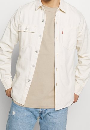Chemise en denim beige clair avec deux poches poitrine, boutons gris et coupe décontractée. Portée par-dessus un t-shirt beige clair et assortie à un jean bleu délavé.