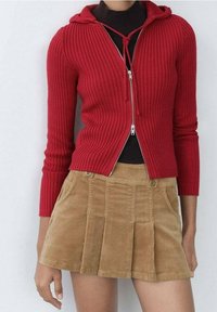 Cardigan rouge côtelé zippé avec capuche, associé à une jupe en velours côtelé beige avec plis et poches avant, soulignée par des détails à boutons.