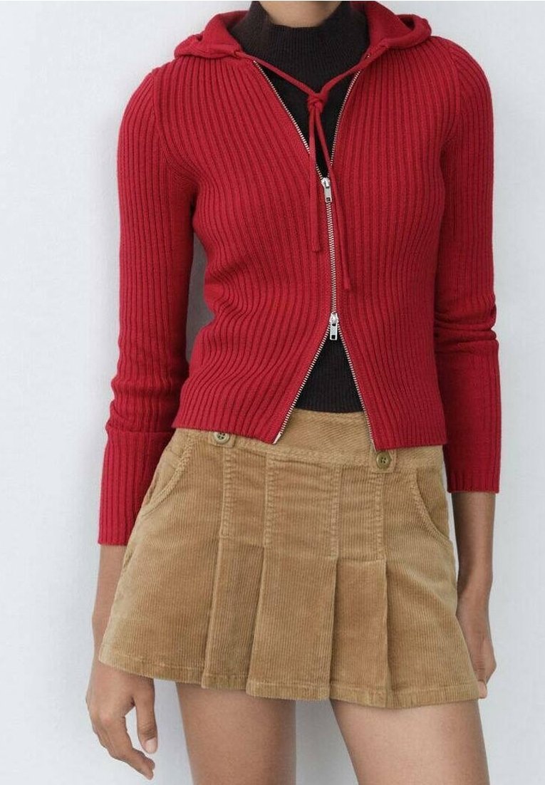 Cardigan rouge côtelé zippé avec capuche, associé à une jupe en velours côtelé beige avec plis et poches avant, soulignée par des détails à boutons.