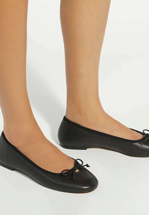Pieds portant des ballerines en cuir noir avec de petits nœuds, sur fond blanc.