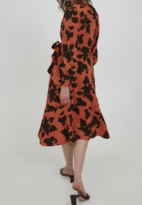 Robe couleur rouille avec un motif de feuilles noires, manches longues, ceinture à la taille et ourlet en tiers. Tissu lisse avec un design fluide. Portée avec des sandales noires.