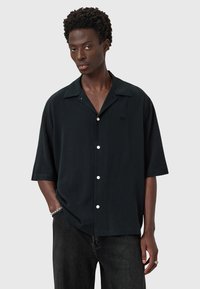 Chemise noire à manches courtes avec col, fermeture à boutons et détail de logo subtil. Le matériau semble lisse avec une coupe décontractée. Associée à un jean foncé.