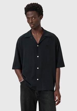 Sort kortærmet skjorte med krave, knappet fortelt og diskret logo detalje. Materialet virker glat med en afslappet pasform. Parret med mørk denim.